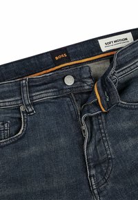 Tmavě modré džíny z denimu se zipovým zapínáním, kovovým knoflíkem a kontrastním oranžovým prošíváním. V pase je značková etiketa 'BOSS' a štítek 'SOFT MOTION'.