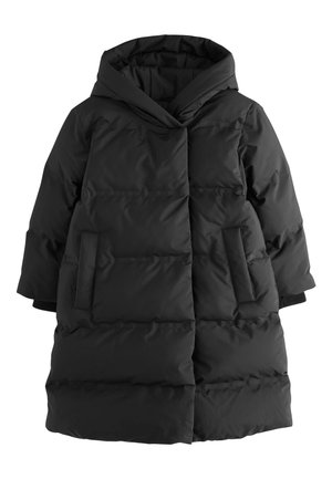 Dijete nosi dugu crnu puhastu jaknu s kapuljačom, sive hulahopke i crne sjajne cipele, stoji ispred jednostavne bijele pozadine.