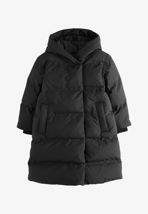 Dijete nosi dugu crnu puhastu jaknu s kapuljačom, sive hulahopke i crne sjajne cipele, stoji ispred jednostavne bijele pozadine.