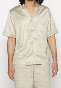 Beige saténový pyžamový set s krátkým rukávem na knoflíky a odpovídajícími kalhotami, zdobený jemným vzorem listů a lemováním.