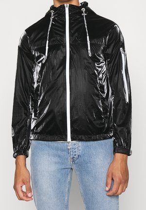Mann trägt eine glänzende schwarze Kapuzenjacke mit weißem Reißverschluss und Kordeln, kombiniert mit hellblauen Jeans, steht vor einem schlichten Hintergrund.