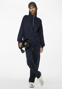 Marineblauwe sportieve zip-up sweatshirt met geribbelde manchetten, gecombineerd met bijpassende joggers. Draagt een zwarte pet en houdt een kleine zwarte tas vast.
