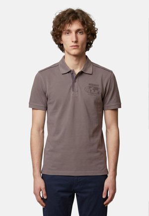 Jeune homme aux cheveux bouclés portant un polo marron avec un petit logo globe sur la poitrine et un pantalon bleu foncé, debout sur un fond blanc.