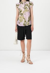 Blouse fleurie à manches courtes, rose et verte, avec des épaules volantes. Shorts noirs avec une ceinture à nouer. Talons beiges à lanières et un petit sac à main blanc.