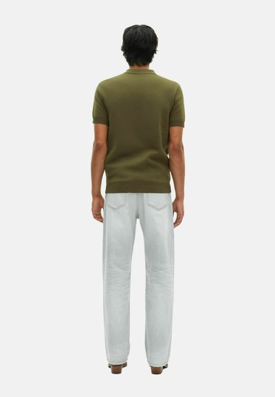 Mand, der står med ryggen til, iført en olivengrøn T-shirt med korte ærmer og lyseblå denim jeans med brune sko, foran en hvid baggrund.