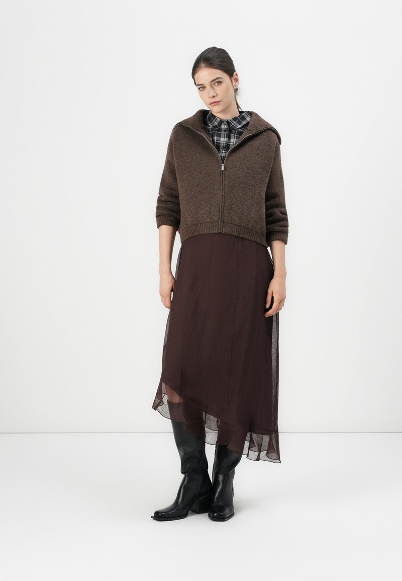 Bruin gebreid cropped vest met een rits, gecombineerd met een donkerbruin doorschijnend asymmetrisch jurk en kniehoge zwarte leren laarzen.