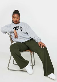 Grå sweatshirt med svart tryck, i kombination med lösa olivgröna byxor och vita sneakers. Modell sitter på en metallstol med armarna vilande.