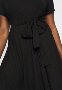 Robe noire à manches courtes avec un détail de taille noué. Un tissu doux et texturé tombe pour former une silhouette fluide.