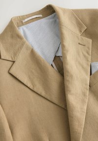 Hele pruun linane bleiser, millel on nurgeline krae ja triibuline puuvillane vooder. Omab minimalistlikku disaini ja peeneid õmblusdetails.