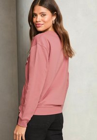 Sudadera de color rosa claro con puños y dobladillo acanalados, que presenta un diseño sencillo. La tela parece suave y tiene un corte relajado.