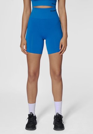 Kobieta stojąca ubrana w niebieskie, wysokie spodenki rowerowe, dopasowany krótki top, białe skarpetki i czarne trampki na jednolitym szarym tle.