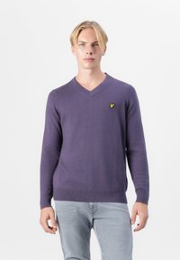Pull violet en V fabriqué en tissu doux, avec des manches longues et un petit logo jaune sur la poitrine. Ourlet et poignets côtes.