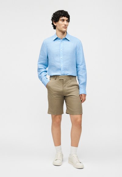 Άνδρας στέκεται φορώντας ανοιχτό μπλε πουκάμισο με μακριά μανίκια και κουμπιά, μπεζ σορτς, λευκές κάλτσες και λευκά αθλητικά παπούτσια μπροστά σε απλό φόντο.