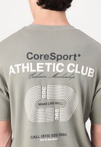 Grå bomulds T-shirt med hvid og sort tekst. Grafikdesignet inkluderer "CoreSport", "ATHLETIC CLUB" og et bane-lignende nummermønster.