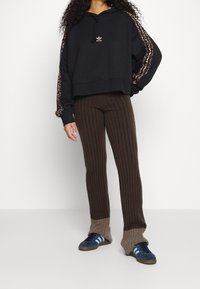 Felpa nera cropped con cappuccio e coulisse, con logo dorato. Pantaloni marroni a coste con polsini di un marrone chiaro a contrasto. Sneakers blu con tre strisce.