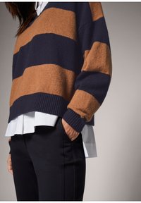 Pull rayé marron et marine avec une texture douce, des poignets côtes et une chemise blanche superposée en dessous, associé à un pantalon sombre.