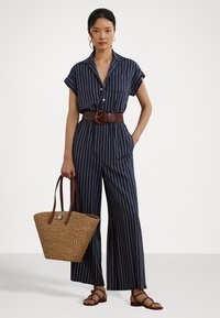 Marine gestreepte jumpsuit met korte mouwen, een kraag en een bruine riem. Model houdt een geweven schoudertas vast en draagt bruine sandalen met bandjes.