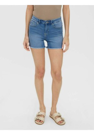 Personne portant un short en jean bleu avec des ourlets effilochés, un haut beige et des sandales beiges, debout sur un fond blanc.