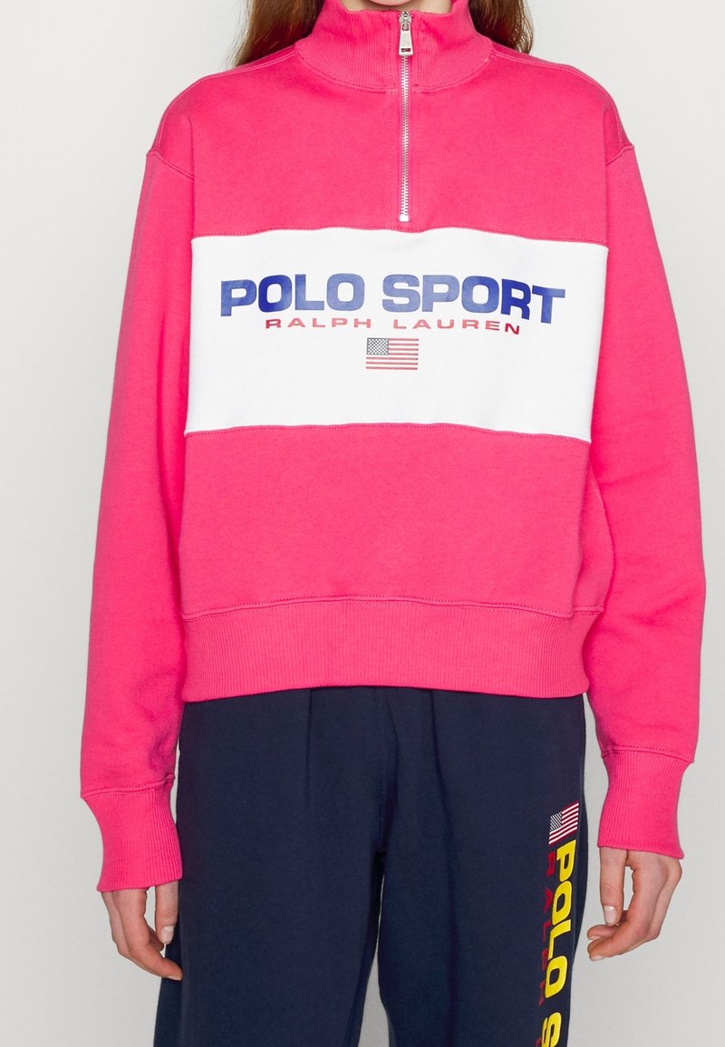 Personne portant un sweat-shirt rose à zip quart avec "Polo Sport Ralph Lauren" et un drapeau américain sur la poitrine. Pantalon bleu marine avec un texte jaune visible.