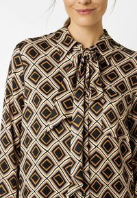 Frau, die eine geometrisch gemusterte Bluse mit schwarzen, beigen und braunen Rauten und einer gebundenen Schleife am Kragen trägt.