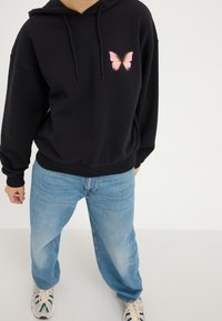 Sudadera negra con un gráfico de mariposa rosa y naranja en el lado izquierdo del pecho, combinada con jeans sueltos de color azul claro y zapatillas.