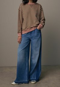 Donna che indossa un maglione a righe rosa e oliva infilato in un paio di jeans a gamba larga blu, con sneakers argentate, in piedi con una mano in tasca.