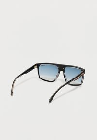 Sončna očala Carrera s črnimi okvirji in modrimi gradientnimi lečami, ki počivajo na belem površju, nagnjena tako, da prikaže podrobnosti okvirja in ročajev.