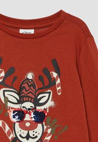 Rdzawy t-shirt z kreskówkowym reniferem w kolorowej zimowej czapce i czerwono-niebieskich okularach przeciwsłonecznych z cekinami, otoczony cukrowymi laskami i wykrzyknikami.
