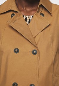 Hnědý dvouřadový kabát s černými knoflíky nošený přes černý a béžový vzorovaný top na tmavé pleti.