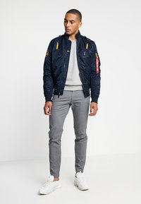 Navy bomber jacket with quilted texture, yellow zippers, and patches, paired with grey slim-fit trousers and white sneakers.