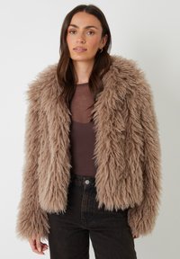 Faux fur-jakke i lys brun med en pelslignende tekstur, der har en bred krave og lange ærmer, sammen med en mørk top og jeans.