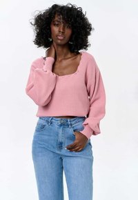 Roze gebreide trui met een brede ronde halslijn en geribde manchetten, gecombineerd met lichtblauwe hoge taille jeans. Textuur van de stof is zichtbaar.