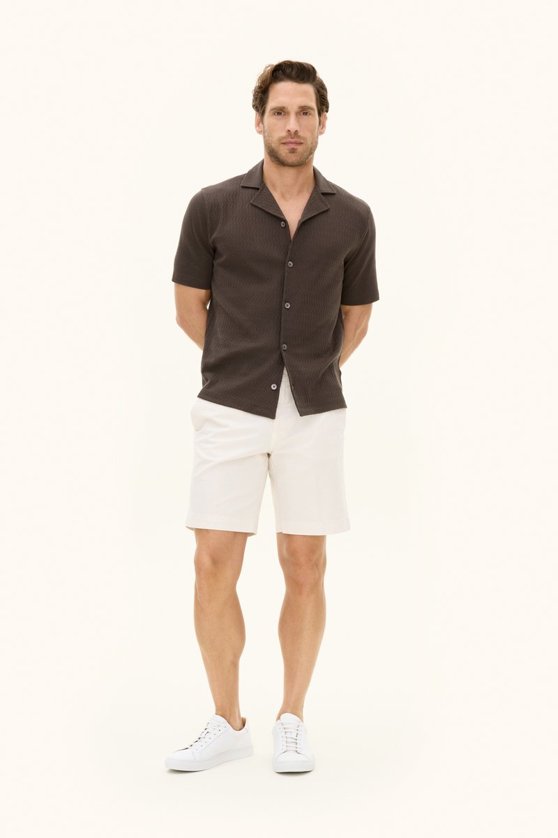 Uomo in piedi con le mani dietro la schiena, indossa una camicia a maniche corte marrone scuro, pantaloncini bianchi e scarpe da ginnastica bianche su uno sfondo semplice.