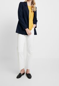 Marineblazer über einem gelben Oberteil, kombiniert mit weißen, geraden Hosen und schwarzen Loafers. Glatte Texturen und taillierte Passform.