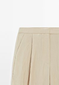 Pantalon beige taille haute avec poches latérales et plis devant, confectionné dans un tissu léger légèrement texturé.