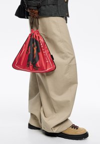 Czerwona torba z troczkami z grafiką, wykonana z lekkiego materiału. Na zdjęciu widoczne są beżowe, luźne spodnie oraz brązowe obuwie na sznurówki.
