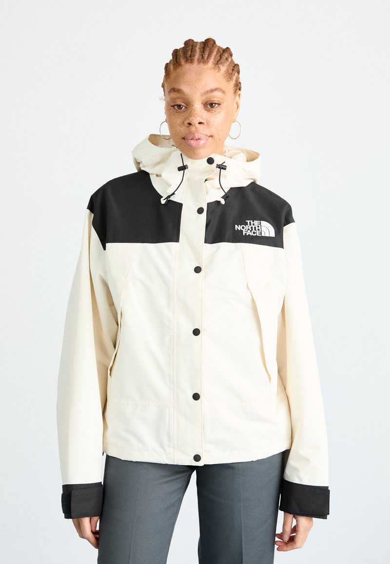 Cream and black waterproof jacket with a hood, featuring a full zip closure and front pockets, made of textured fabric, logo on chest.