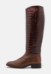 Bottes hautes en cuir marron avec un dos brillant à motif crocodile. Caractérisées par un bout arrondi et un petit talon bloc, rehaussées de coutures blanches.