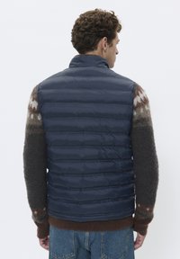 Marineblauer Steppwesten mit horizontalem Steppmuster, hohem Kragen, kombiniert mit einem mehrfarbigen Muster-Strickpullover. Weiches, isolierendes Material.