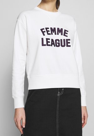 Kvinde iført en hvid sweatshirt med teksten "FEMME LEAGUE" og et sort skørt med hvide syninger.