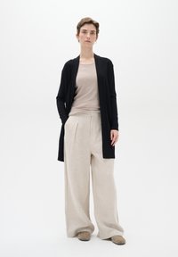 Zwarte lange cardigan over een beige top, gecombineerd met lichtbeige wijde broek. De outfit heeft zachte texturen en neutrale kleuren.
