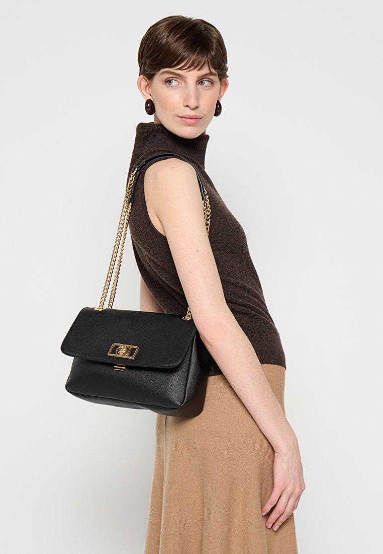 Femme portant un col roulé sans manches marron et une jupe beige, avec un sac à main en cuir noir à bandoulière chaîne dorée sur l'épaule.