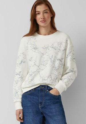 Młoda kobieta ubraną w białą bluzę z czarnymi liniowymi kwiatowymi wzorami, w połączeniu z niebieskimi dżinsami, pozująca z jedną ręką w kieszeni.