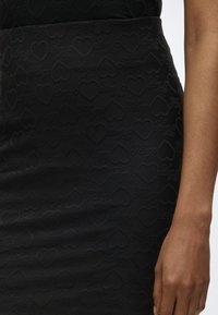 Μαύρη υφασμάτινη φούστα με ανάγλυφα σχήματα καρδιάς και καλλιγραφικό κείμενο "Flying", φορεμένη από άτομο με εμφανές χέρι και βραχίονα.