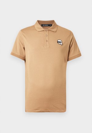 Polo color beige realizzata in cotone, con una patta a tre bottoni e un logo bianco e nero sul lato sinistro del petto. Maniche corte, texture liscia.
