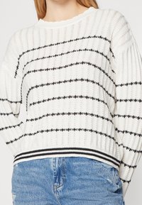 Biały sweter dziany z czarnymi, poziomymi paskami. Posiada prążkowany dekolt i mankiety oraz teksturowany materiał i luźny krój.