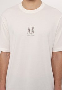 Balta medvilninė marškinėliai su pilku siuvinėjimu "A|X" logotipu ir tekstu "ARMANI EXCHANGE" apačioje, su klasikiniais apykaklės ir trumpomis rankovėmis.