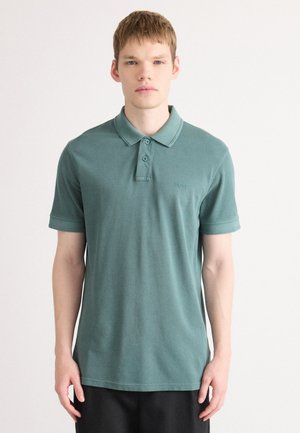 Tânăr cu păr scurt, brunet deschis, purtând un tricou polo verde deschis cu mânecă scurtă, stând în fața unui fundal alb simplu.
