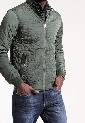 Uomo che indossa giacca trapuntata verde con zip e tasche con bottoni a pressione e jeans blu, mani nelle tasche, in piedi davanti a sfondo bianco.