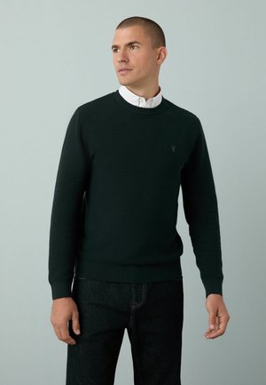 Pull en laine vert foncé avec un design en tricot texturé, col rond et poignets côtelés. Porté par-dessus une chemise blanche à col et associé à un jean foncé.